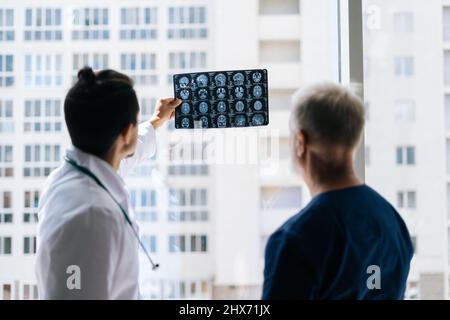 Rückansicht eines erwachsenen Arztes und eines jungen Radiologen, die gemeinsam die Krankengeschichte eines Patienten mit einer MRT-Untersuchung des Kopfes in der Nähe des Fensters besprechen. Stockfoto