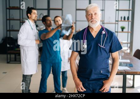 Porträt eines reifen älteren Chirurgen in blauer Uniform, der im Büro eines medizinischen Meetings steht und auf die Kamera schaut. Stockfoto