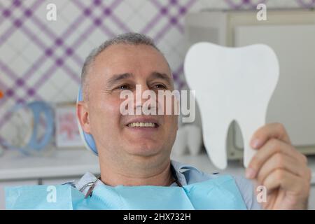 Ein Mann mittleren Alters sitzt auf dem Zahnstuhl und nutzt einen Spiegel, um das Ergebnis der Stomatologie zu sehen, die durchgeführt wurde Stockfoto