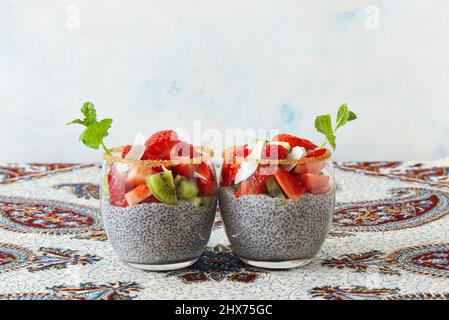 Blick aus der unteren Ecke auf Chia Pudding Dessert mit Sommerbeeren, Erdbeeren, Kiwi und Kokosnussscheiben, verziert mit einem Minzzweig. Keine Personen. Stockfoto