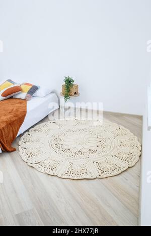 Niedliches selbstgemachtes Strickmuster aus dicken Fäden von Hand: Ein handgefertigter Teppich aus beigen Fäden liegt im Schlafzimmer neben dem Bett, die Kunst des Strickens Stockfoto