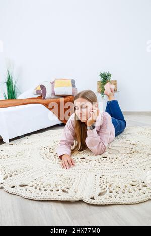 Eine Frau liegt auf einem Teppich in der Nähe des Bettes, ein selbstgemachter Strickteppich aus dicken Fäden, von Hand gefertigt: Ein handgefertigter Teppich aus beigen Fäden liegt auf dem Teppich Stockfoto