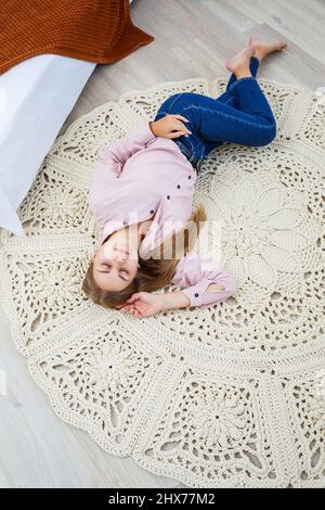 Eine Frau liegt auf einem Teppich in der Nähe des Bettes, ein selbstgemachter Strickteppich aus dicken Fäden, von Hand gefertigt: Ein handgefertigter Teppich aus beigen Fäden liegt auf dem Teppich Stockfoto
