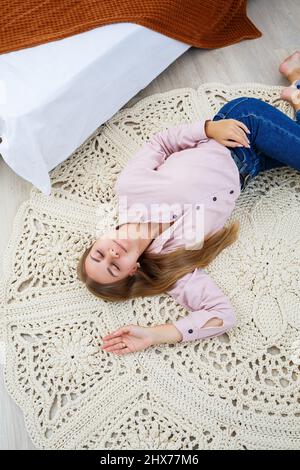 Eine Frau liegt auf einem Teppich in der Nähe des Bettes, ein selbstgemachter Strickteppich aus dicken Fäden, von Hand gefertigt: Ein handgefertigter Teppich aus beigen Fäden liegt auf dem Teppich Stockfoto