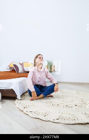 Eine Frau liegt auf einem Teppich in der Nähe des Bettes, ein selbstgemachter Strickteppich aus dicken Fäden, von Hand gefertigt: Ein handgefertigter Teppich aus beigen Fäden liegt auf dem Teppich Stockfoto