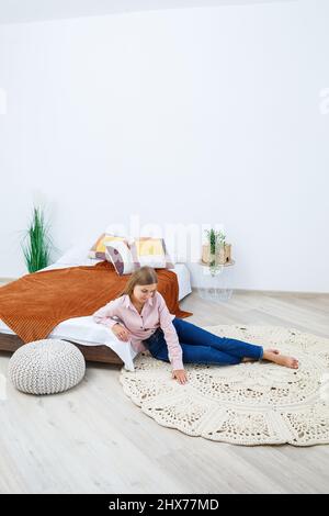 Eine Frau liegt auf einem Teppich in der Nähe des Bettes, ein selbstgemachter Strickteppich aus dicken Fäden, von Hand gefertigt: Ein handgefertigter Teppich aus beigen Fäden liegt auf dem Teppich Stockfoto