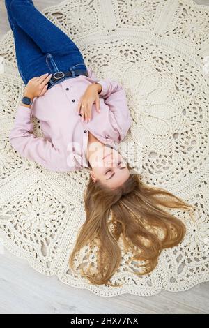 Eine Frau liegt auf einem Teppich in der Nähe des Bettes, ein selbstgemachter Strickteppich aus dicken Fäden, von Hand gefertigt: Ein handgefertigter Teppich aus beigen Fäden liegt auf dem Teppich Stockfoto