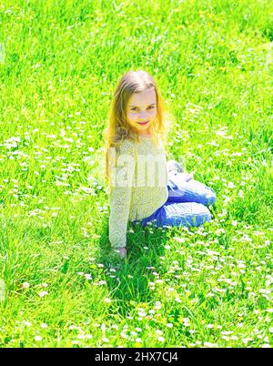 Mädchen sitzt auf Gras auf Grünfläche, grüner Hintergrund. Das Kind genießt das sonnige Frühlingswetter, während es auf der Wiese sitzt. „Heyday“-Konzept. Mädchen auf lächelndem Gesicht Stockfoto