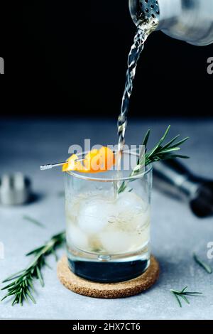 Alkoholcocktail aus dem Shaker in Glas mit Orangenschale Rosmarin und Eis auf Holztisch Stockfoto