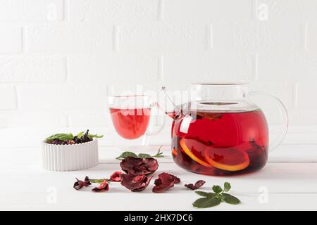 Eine Glas-Teedose mit frisch gemahlenem Hibiskustee und Zitrone an einer weißen Ziegelwand und einer Tasse Tee. Vitamintee zur Gewichtsreduktion Stockfoto