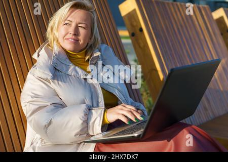 Schöne kaukasische Frau mittleren Alters, die auf einem Laptop auf der Holzbank im Freien sitzt. Stockfoto