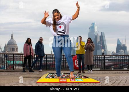 NUR FÜR REDAKTIONELLE VERWENDUNG Mitglieder der Öffentlichkeit sehen die Schauspielerin Jacqueline Jossa, die barfuß über einen aus 30.000 LEGO Steinen gefertigten Pfad am Observation Point in London geht, um Geld für den Red Nose Day 2022 von Comic Relief zu sammeln. Bilddatum: Donnerstag, 10. März 2022. Stockfoto