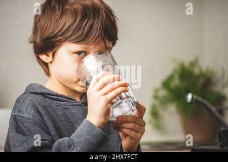 Weißer Junge hält ein Glas Wasser in der Küche in der Nähe des Waschbeckens. Gereinigtes Leitungswasser, das sicher zu trinken ist Stockfoto