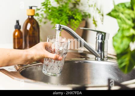 Weibliche Hand hält ein Glas und gießt es mit Leitungswasser in die Küche zu Hause. Bleib hydratisiert und filtrierende Wasser Konzept. Weltwassertag Stockfoto