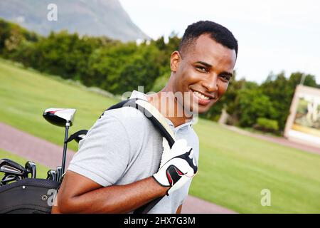 Bereit, auf dem grünen zu erhalten. Porträt eines hübschen jungen Mannes, der mit seiner Tüte Schläger auf einem Golfplatz steht.