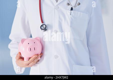 Stellen Sie sicher, für unerwartete medizinische Kosten zu planen. Ausgeschnittene Aufnahme einer nicht erkennbaren Ärztin, die ein Sparschwein hält, während sie im Krankenhaus steht. Stockfoto