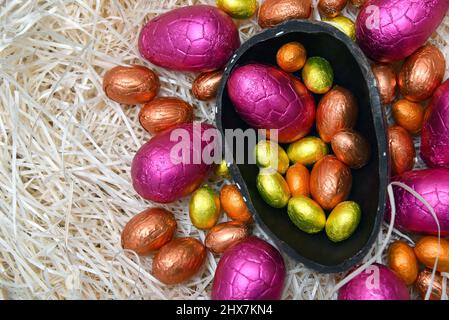 Stapel von bunten, in Folie verpackten Schokoladen-ostereiern in Rosa, Rot, Silber und Gold mit zwei Hälften eines großen braunen dunklen Schokoladeneier. Stockfoto