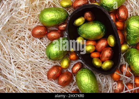 Stapel von bunten, in Folie verpackten Schokoladen-ostereiern in Grün, Orange, Gelb und Gold mit zwei Hälften eines großen braunen dunklen Schokoladeneier. Stockfoto