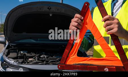 Junger Mann, der ein rotes Dreieck vorbereitet, um andere Verkehrsteilnehmer zu warnen, Autoausfall oder Motorausfall halten an der Landstraße an. Männlicher Fahrer, der in der Nähe eines b steht Stockfoto