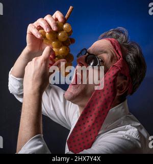 Ein Mann mit einer roten Krawatte auf dem Kopf und einer Sonnenbrille hält einen gefälschten Traubenhaufen, als wolle er ihn essen Stockfoto