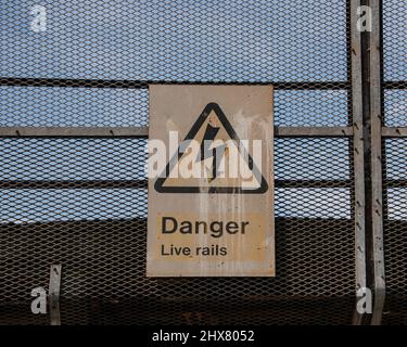 Ein Schild, das an Geländern neben einer Eisenbahnlinie befestigt ist und vor stromführenden Schienen warnt. Stockfoto