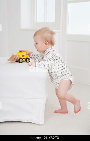 Kleiner Junge, der gegen das Bett steht und mit einem gelben LKW-Spielzeug spielt Stockfoto