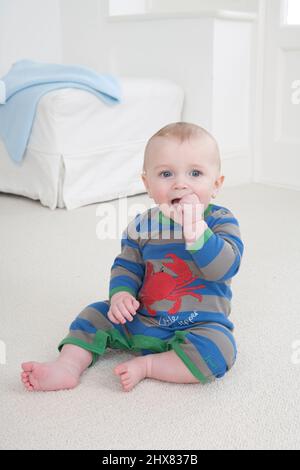 Kleiner Junge, der auf dem Teppich sitzt, mit den Fingern einer Hand im Mund Stockfoto