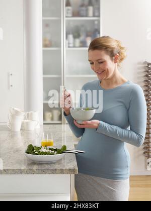 Lächelnde Schwangere, die neben der Küchenarbeitsfläche steht und eine Schüssel mit eisenangereicherten Gemüseblättern hält Stockfoto