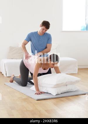 Mann massiert den unteren Rücken seines Schwangeren, während sie während ihrer ersten Wehen auf allen Vieren im Wohnzimmer auf einer Matte kniet Stockfoto