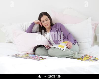 Müde, Schwangere, die sich auf dem Bett entspannen, das von Kissen umgeben ist von Zeitschriften, Vorderansicht Stockfoto