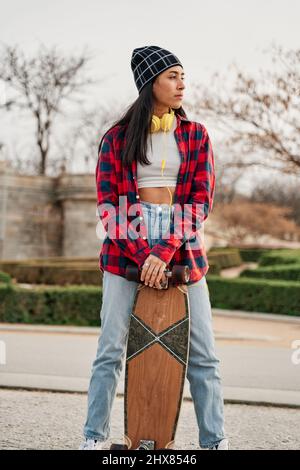 Schöne junge Hipster Frau posiert mit Skateboard. Lateinamerikanische Frau in legerer Kleidung. Stockfoto