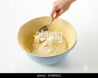 Mehl, Backpulver und gemahlene Mandeln zu einer Mischung aus Eiern, Gießzucker, Butter und Rosenwasser zusammenfalten, um einen mit Rosen und Mandeln bemalten Kuchen zu machen. Verwenden eines großen Metalllöffels in einer blauen Rührschüssel. Stockfoto