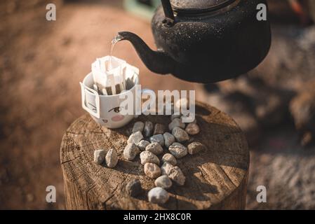 Der Prozess der Herstellung eines Tropfkaffees mit dem gekochten Wasser aus einem alten schwarzen Topf auf dem Kamin beim Camping oder Wandern. Stockfoto