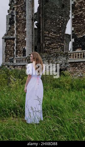 Geisterhaft blasse junge Frau in weißem Kleid, die neben den Ruinen einer alten normannischen Kirche steht Stockfoto