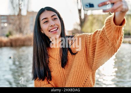 Foto von sorglosen langhaarigen jungen lateinischen Frau nimmt Selfie-Porträt auf dem Handy, trägt gelben bunten Pullover, nutzt Technologie in der Freizeit ti Stockfoto