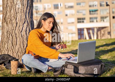Junge Latina mit langen Haaren mit einem Laptop, während sie die Ukulele unter einem Baum in der Stadt spielt. Stockfoto