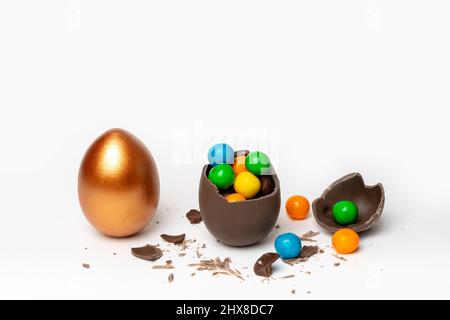 Knackige Schokolade osterei mit bunten kleinen runden Süßigkeiten und Pralinen und ein ganzes bemaltes goldenes Ei auf weißem Hintergrund, Kopierraum. Ostern Stockfoto