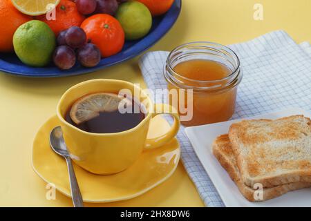 Frühstück - Toast Brot, Tee mit Zitrone, Honig im Glas, Obst auf einem Teller. Und eine schöne gelbe Tischdecke. Stockfoto