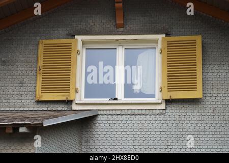 Fenster mit offenen Fensterläden. Weißes Hemd hängt im Fenster. Hausfassade in der Schweiz. Die Fassade ist mit Holzschindeln bedeckt. Stockfoto