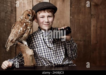 Ornithologie macht so viel Spaß. Kleiner Junge in Retro-Kleidung mit Brille, der ein Fernglas neben einer Eule hält. Stockfoto