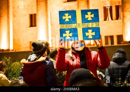 Tiflis, Georgien - 1.. märz 2022: Frau mit georgisch-ukrainischen Gesichtsmaske halten und zeigen handgemachte Gemälde der ukrainischen Flagge mit dem Titel "Ukrainer Stockfoto