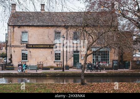 Die Menschen machen eine Pause auf Bänken vor einem traditionellen Pub in dieser verschimmenen Cotswold-Stadt. Stockfoto