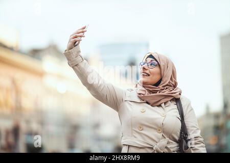 Eine muslimische Frau mittleren Alters trägt einen Hijab mit einem glücklichen Gesicht, das in einer städtischen Umgebung steht und Selfie mit ihrem Smartphone macht. Stockfoto