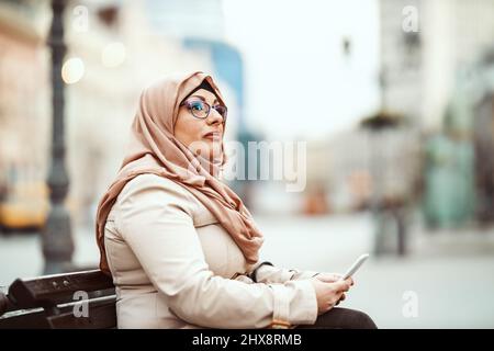 Eine muslimische Frau mittleren Alters, die einen Hijab mit einem glücklichen Gesicht trägt, sitzt auf der Bank in einer städtischen Umgebung und gibt Nachrichten auf ihrem Smartphone ab. Stockfoto