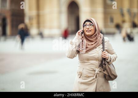 Eine muslimische Geschäftsfrau mittleren Alters trägt einen Hijab mit einem glücklichen Gesicht, das in einer städtischen Umgebung steht und ihr Smartphone anzieht. Stockfoto