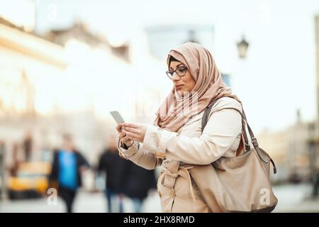 Eine muslimische Frau mittleren Alters trägt einen Hijab mit einem glücklichen Gesicht, das in einer städtischen Umgebung steht und Nachrichten auf ihrem Smartphone gibt. Stockfoto