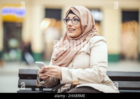 Eine muslimische Frau mittleren Alters, die einen Hijab mit einem glücklichen Gesicht trägt, sitzt auf der Bank in einer städtischen Umgebung und gibt Nachrichten auf ihrem Smartphone ab. Stockfoto