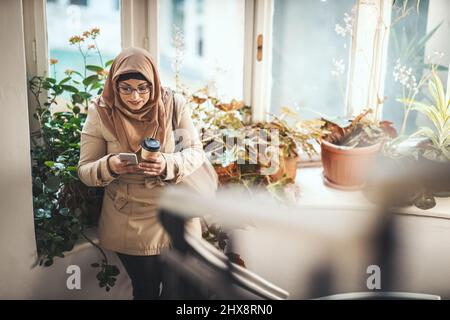 Eine muslimische Frau mittleren Alters, die einen Hijab mit einem glücklichen Gesicht trägt, steht am Fenster und tippt Nachrichten auf ihrem Smartphone. Stockfoto