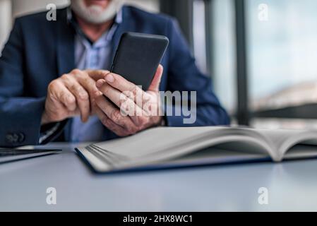 Ein gesichtsloser, älterer Mann hält das Smartphone, um Nachrichten und E-Mails abzulesen, während er in einem modernen, hellen Büro sitzt. Konzentrieren Sie sich auf Hand und Telefon. Stockfoto