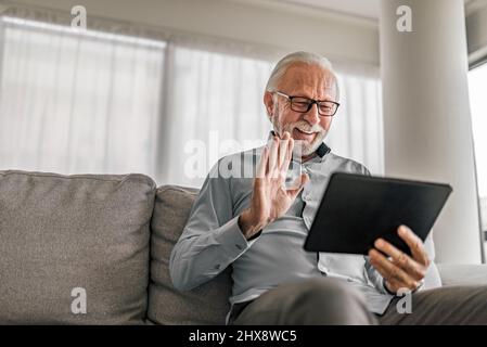 Fröhlicher älterer Mann mit Videoanruf auf Tablet sitzt auf der Couch zu Hause älterer Mann trägt eine Brille und bleibt mit Freunden und Familie in Kontakt Stockfoto